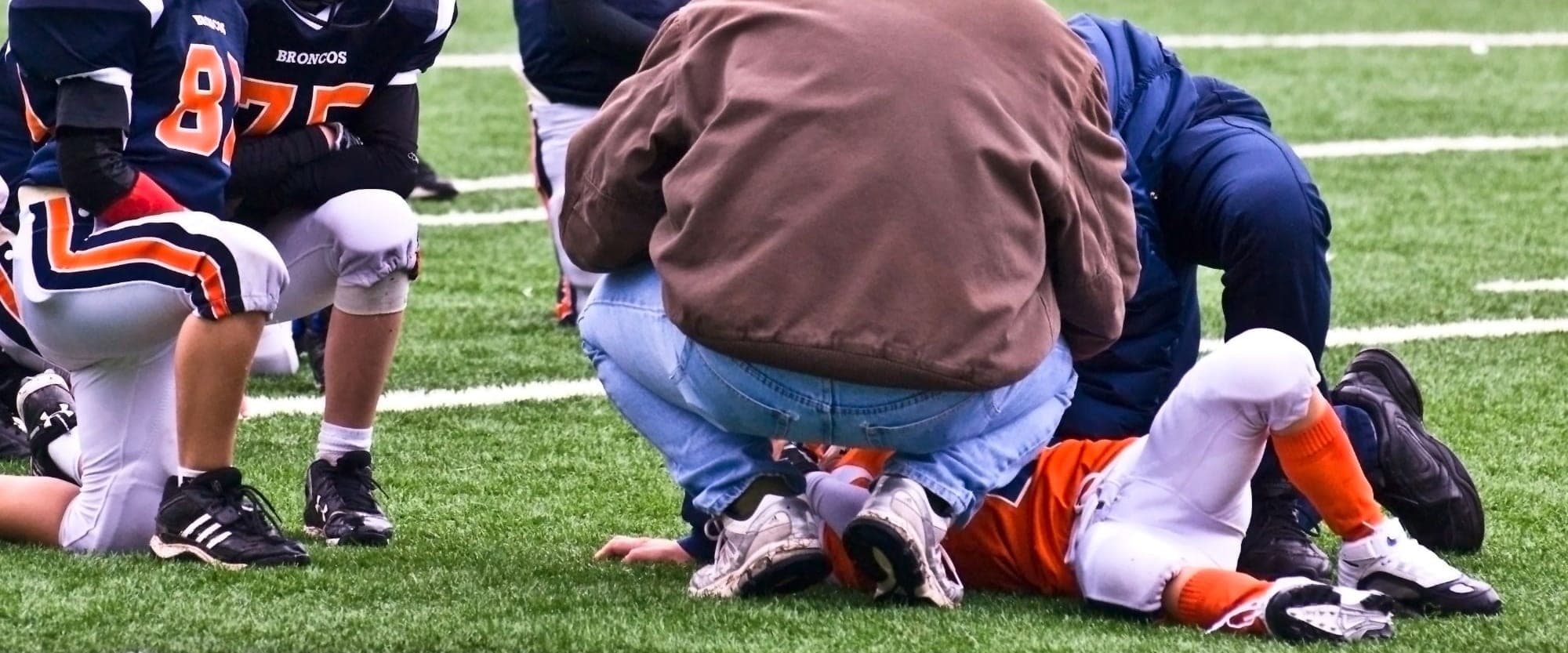 A youth football player lying on the field while two adults attend to him and his teammates kneel nearby